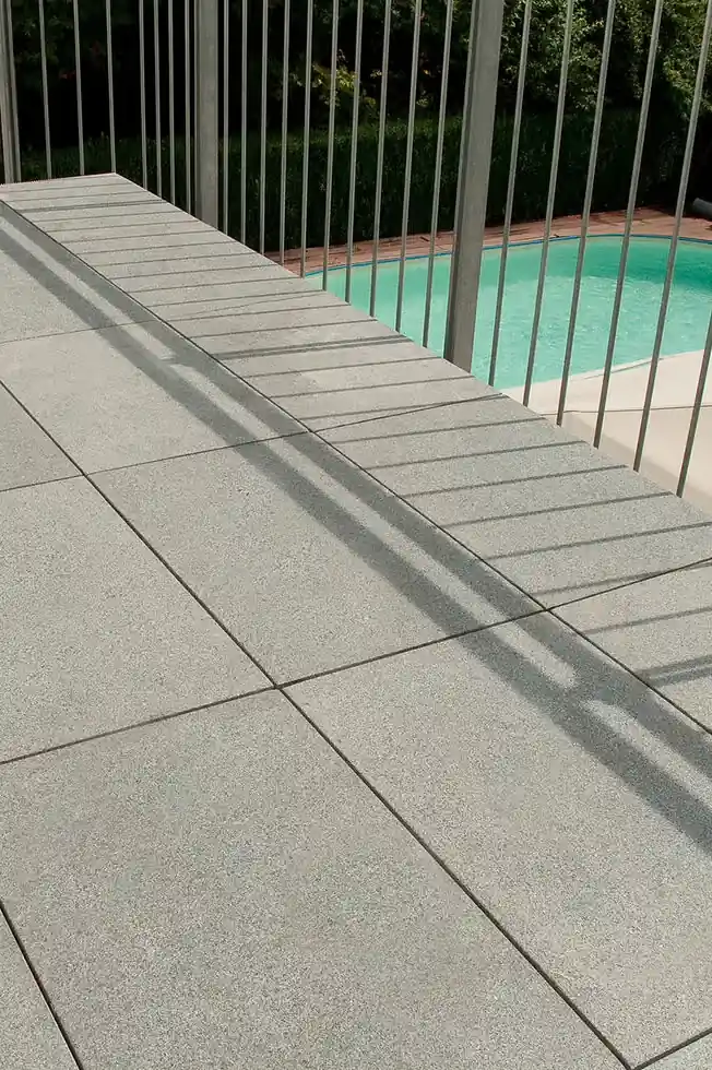 Light gray tiled patio area with vertical metal railing casting shadows and a swimming pool beyond. PLATTENFIX Fugenkreuz für gleichmäßige Fugenbilder bei Terrassenplatten