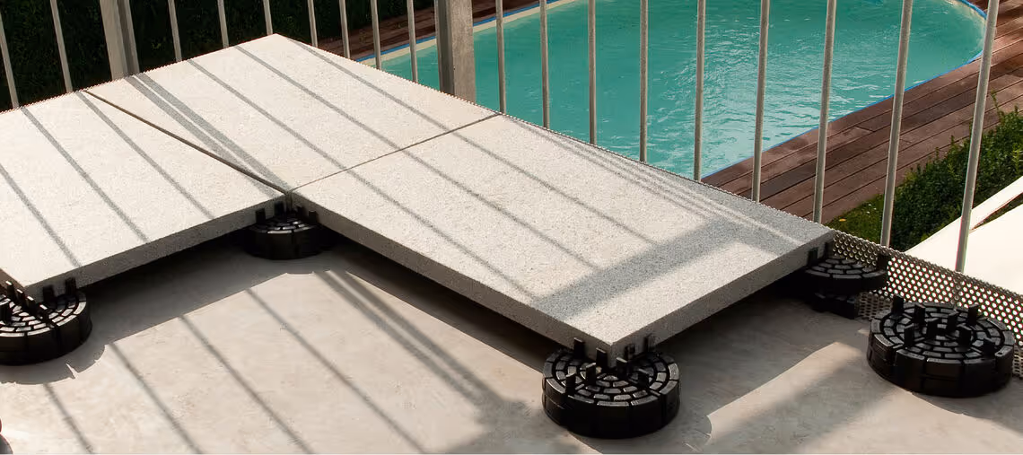 Stone tiles elevated on adjustable black pedestals near a pool with metal railing shadows cast over them.