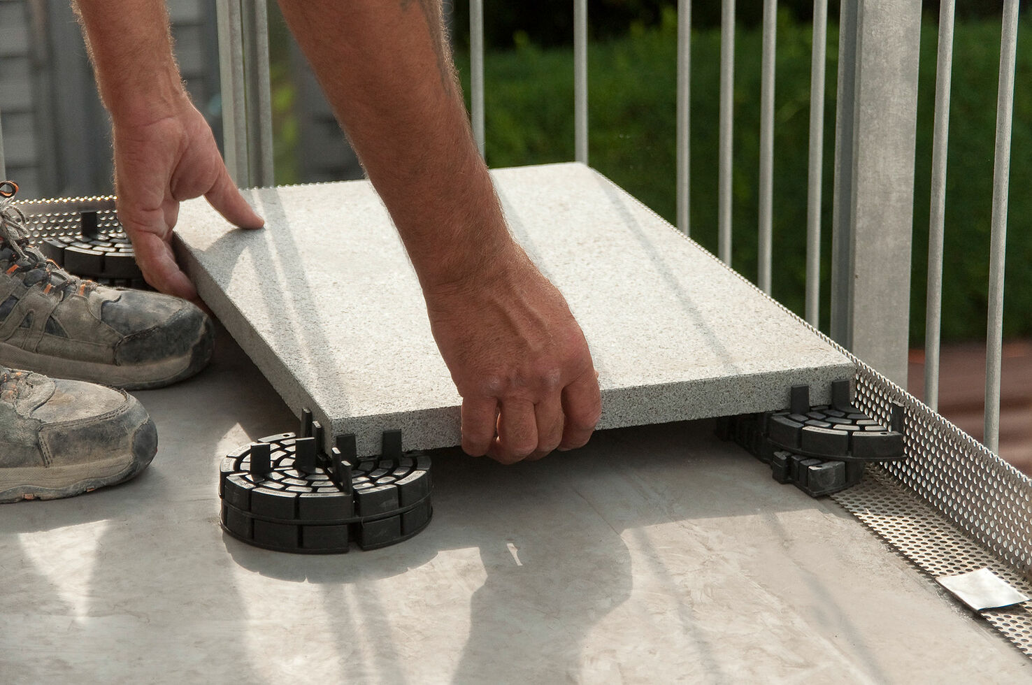 Close-up of hands placing a large stone tile on adjustable black plastic supports on an outdoor balcony floor.