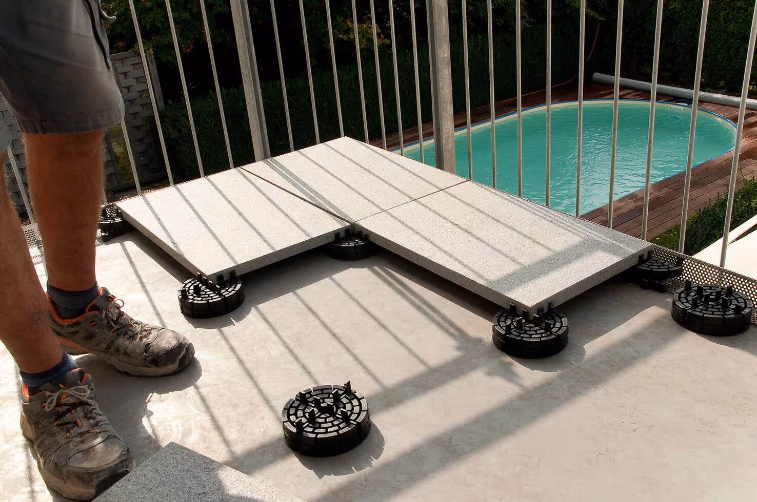 Person installing large gray tiles on adjustable black supports on a balcony next to a pool. PLATTENFIX Rasenfugenkreuz für ökologische Auffahrten und Versickerung