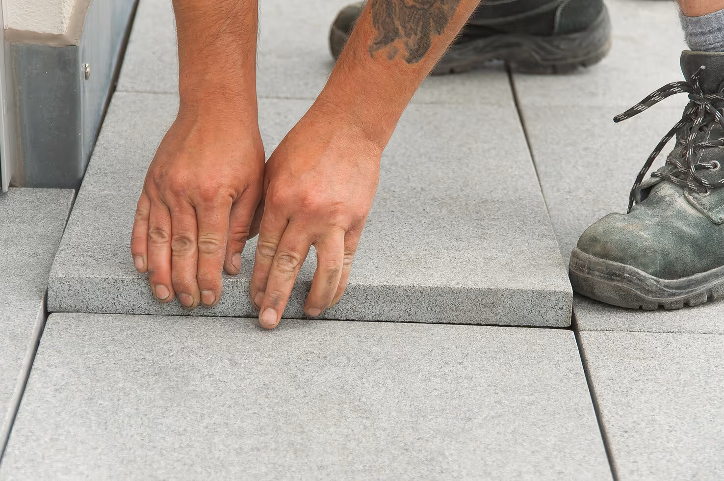 Hands placing a gray paving stone on the ground next to already laid stones, with a boot visible.PLATTENFIX MAXI Plattenlager für unebene Untergründe und Dachterrassen