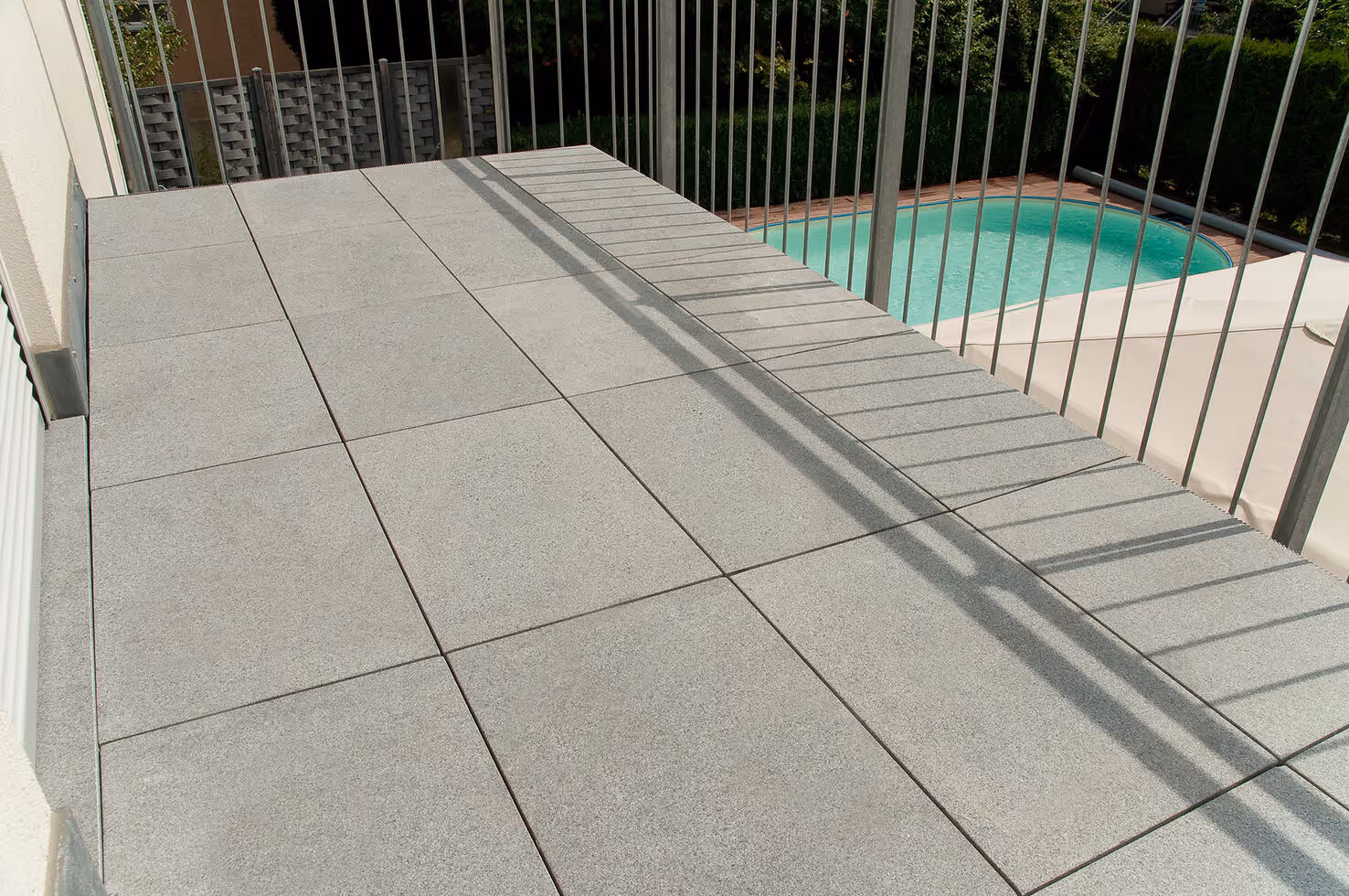Balcony with large light gray square tiles and metal railing overlooking a swimming pool and green shrubs.